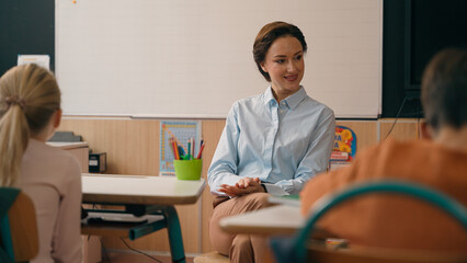 Obraz premium Little children kids classmates pupils students sitting at desks tables in classroom listening Caucasian woman teacher in elementary school teach class studying education explain task lesson teaching