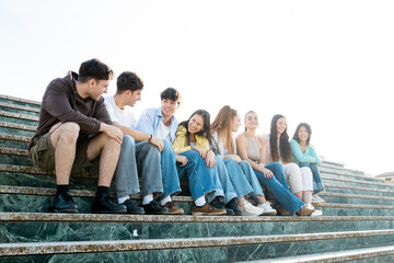 Side photo of a group of 8 young people of different ethnicities between 18 and 25 years old having fun in a city.The millennials are sitting on some steps while chatting.