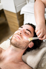 Obraz premium Vertical photo of a handsome bearded man in his 30s and 40s is inside a spa.The male is lying on a table while undergoing a facial massage with a brush.Massages with brushes and oil.