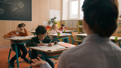 Unrecognizable woman teacher tutor ask children lesson check knowledge of pupils schoolboy little African American boy raise hand answer clever smart child kid son reading in book at class school