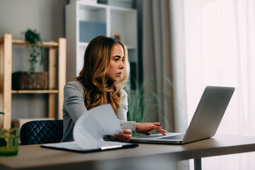 Young woman working at home office using laptop and analyzing documents