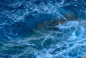 Waves crashing over sunlit rocks in a turquoise sea on a serene afternoon.