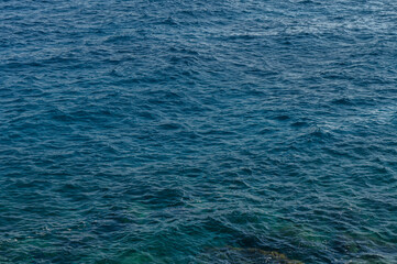 Obraz premium Gentle waves lap against the rocky shoreline under a clear blue sky at midday by the sea.