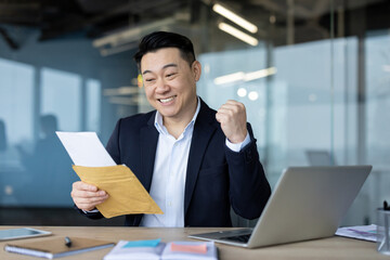 Fototapeta premium Asian young male businessman sitting in the office at the desk, holding an open envelope in his hands, reading a reminder letter and happy with the news, showing a victory gesture with a yes hand