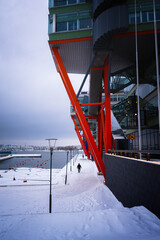 Man walking on snowy path during winter by water next to a modern building with red structures
