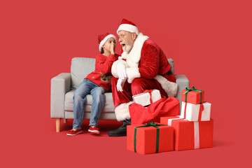 Shocked Santa Claus and cute little girl with gift boxes telling her wish on sofa against red...