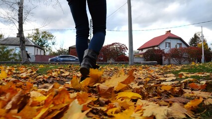 Legs of young woman stepping on yellow foliage at park. Female feet in boots going on fallen maple leaves at parkland. Girl walking at parkland at autumn season on cloudy day. Close up Slow motion