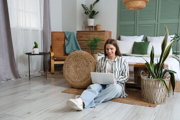 Pretty young woman with laptop sitting near bed in bedroom