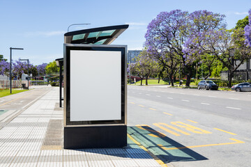 Vertical blank billboard at a bus stop shelter in Buenos Aires, Argentina with jacaranda trees blooming along Libertador avenue in spring. Copy space poster on a lightbox for displaying ad content