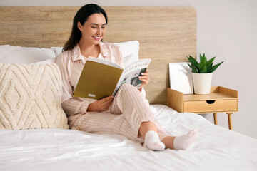 Young woman in pajamas reading magazine on bed at home