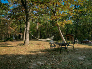 Autumn Backyard Hammock. A hammock supended between two trees in a  forested backyard with a table and chairs in the foreground during the leaf color change of Autumn.