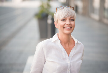 European happy blond adult woman looking to camera. Smiling face of beautiful mid aged female person dressed in white summer shirt. Outdoors