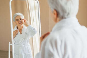 Obraz premium Love yourself. Beautiful old woman enjoying mirror reflection. Senior older lady spending morning in bathroom looking at mirror confident happy. Woman doing daily morning beauty routine. Self love