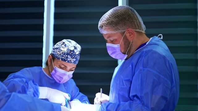 Attentive female surgeon carefully uses electric scissors. Bearded doctor assisting at operation on thyroid.