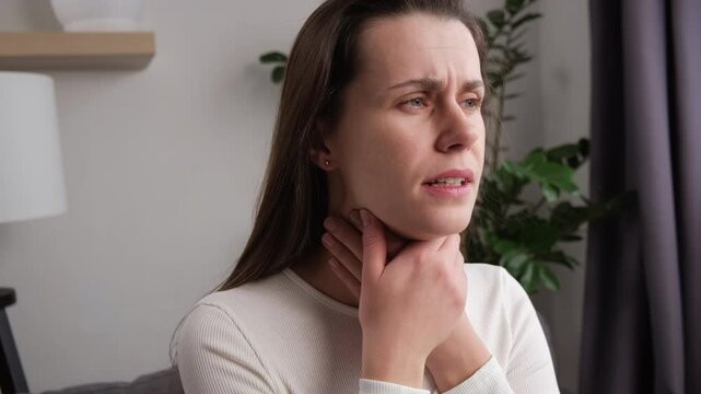 Sickness in inflaming young woman use hands check self touch at sore throat, pain thyroid gland on neck or disease reflux, acid of suffer girl sitting on couch at home. Medical and healthcare concept