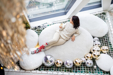 Happy Asian woman sitting in snowy window inside of festive room with christmas tree lights and gifts. Atmospheric christmas eve at home. Merry Christmas!