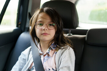 Little girl with glasses sits fastened with seat belt in car. Concept of children's travel, safety, activity, fun and joy