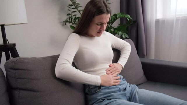 Sick young woman in pain holding belly stomach feeling hurt abdomen ache gastritis pancreatitis symptoms. Upset female sitting on couch suffering from indigestion flatulence abdominal problem concept