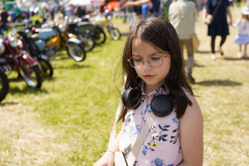 Fototapeta premium Little girl in glasses and headphones having fun against the background of old cars and motorcycles. Concept of children's travel, activity, fun and getting new impressions