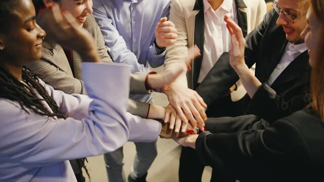 A close-up of joyful, diverse business professionals holding hands in unity, showing teamwork and a strong sense of camaraderie in a modern loft office.