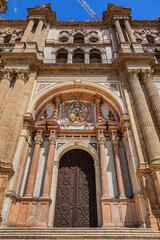 Fragments of majestic facade of Malaga Cathedral (1782), Baroque and Renaissance architecture with grand columns, detailed carvings, and archways under a clear blue sky. Malaga, Andalusia, Spain.