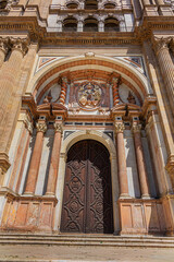 Fragments of majestic facade of Malaga Cathedral (1782), Baroque and Renaissance architecture with grand columns, detailed carvings, and archways under a clear blue sky. Malaga, Andalusia, Spain.