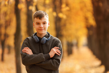 A teenager stands against the backdrop of an autumn park, arms crossed over his chest. Headphones rest casually around his neck, and he wears a slight smile, gazing directly at the camera. 