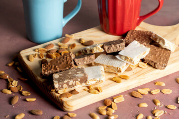 Close-up of various nougat and chocolate bars with almonds and peanuts on a wooden board, accompanied by red and blue mugs in a rustic setup.
