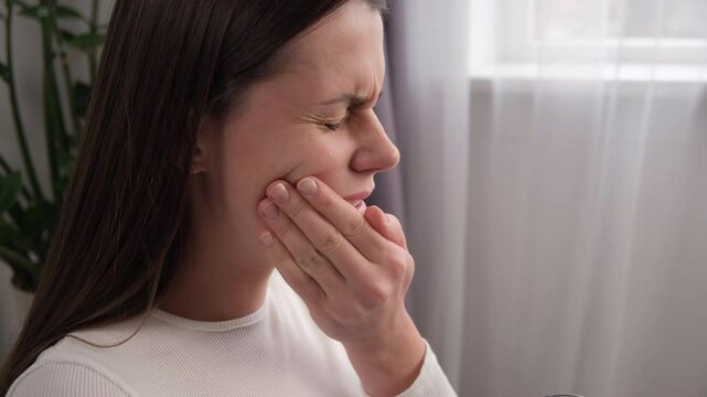 Close up of young woman feel terrible toothache after drink cold water. Female sitting on sofa at home touching cheek, feel hurt and suffering from sensitive tooth ache. Pain and cavities concept