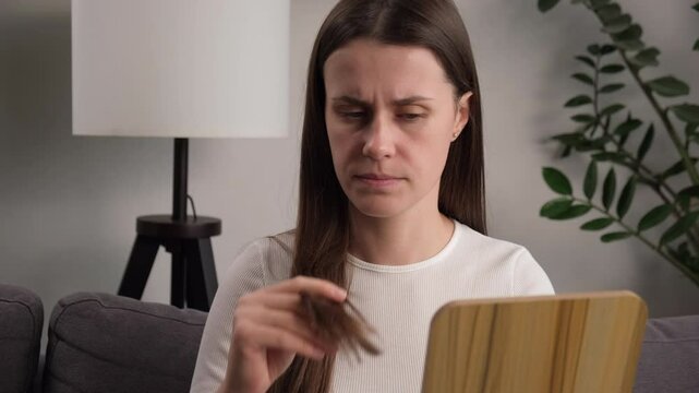 Frustrated worry young woman holding splitting ends, messy unbrushed dry hair and frizzy with disheveled hair looking in mirror sitting on couch at home. Damaged hair. Health care of beauty concept