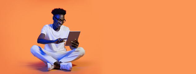 A young black guy sits cross-legged on a vibrant orange background, engaged with a tablet device. The person is wearing a white t-shirt and jeans, focused on their screen.