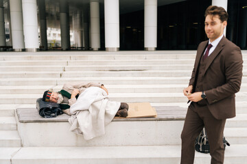 Portrait of successful businessman looking at camera holding money while standing near homeless person sleeping on stairs covered with blanket in front of modern urban building.