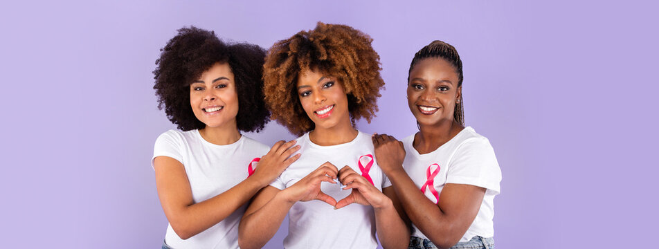 Oncology Awareness. Trio Of Positive African American Ladies With Pink Breast Cancer Ribbons Hugging, Woman Shaping Fingers Like Heart Over Purple Studio Background. Support Group, Copy Space