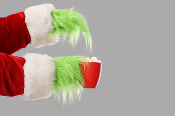 Green hairy hands of creature in Santa costume pointing at cup with cocoa on grey background