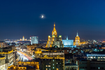 Obraz premium The skyline of Moscow sparkles with vibrant lights from historical buildings under a crescent moon, showcasing the city's beauty at night.