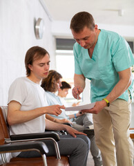 Fototapeta premium Professional doctor showing papers while explaining results of medical examination to young male patient in lobby of clinic