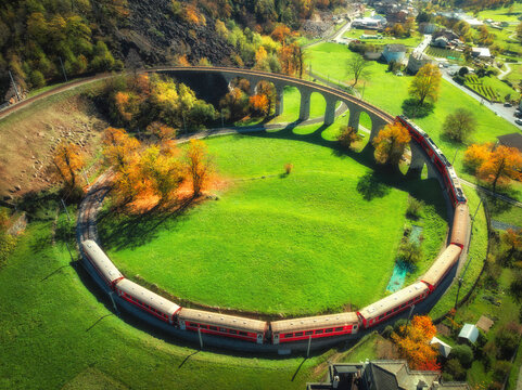 Aerial view of red train on Kreisviadukt in alpine mountains at sunset in autumn. Bernina Express, Switzerland. Top view of train, railroad, green grass , colorful trees in fall. Brusio spiral viaduct