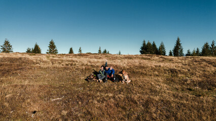 Obraz premium A young couple travels in the mountains with two dogs. A man, a young woman and their Australian and German Shepherd sit in a clearing in the Komovi Mountains, Montenegro. Aerial view.
