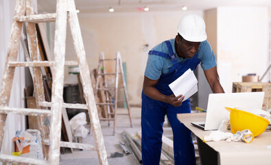 Fototapeta premium African american civil engineer, standing on a construction site with drawings in his hands, checks the house reconstruction ..plan on a laptop
