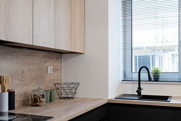 Modern kitchen interior with black kitchen sink with faucet near window with blinds. Wooden counter and cabinets in contemporary room. Design home.