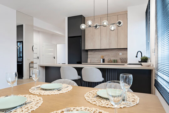 Modern dining room interior with kitchen with wooden counter and dining table with empty plate and glass. Design furniture with stylish glass lapm over the table. Real estate.	