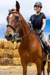 Obraz premium Smiling female rider on a brown horse near hay bales