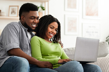 Fototapeta premium Cheerful Black Couple Using Laptop At Home, Watching Movies Or Ordering Food Delivery, Empty Space