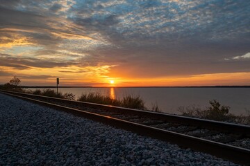 Railroad tracks running along the shore of a lake with a fiery orange, blue, and yellow sunet glowing in the background.