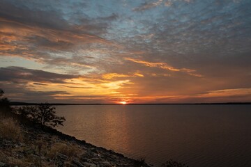 Fiery orange and yellow clouds filling the sky over the calm water of a lake as the sun sets on the distant horizon.