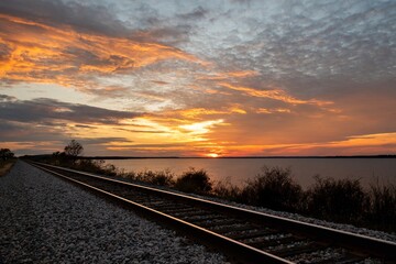 Railroad tracks leading off into the distance along the shore of a lake as the sunset fills the sky with fiery yellow and orange clouds.