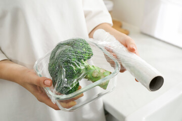 Woman putting plastic food wrap over glass container with broccoli in kitchen, closeup