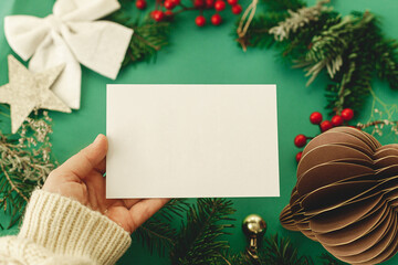 Hand holding empty christmas card on green background with fir branches, red berries and paper...