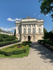 Front view of an elegant historical building with classic architecture surrounded by a manicured garden under a clear blue sky. Ideal for themes of history, architecture, and travel.