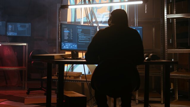 Hacker arrives in messy base of operations with neon lights and graffiti drawings sprayed on walls. Cybercriminal using ghetto hideout as HQ to commit illegal activities using computer, camera A